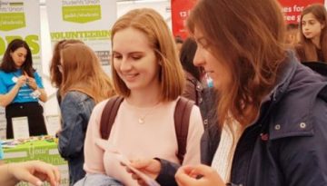 BAM Agency At a bustling job fair, people engage around vibrant stalls. A person in a blue shirt hands out information to attentive attendees clutching marketing pamphlets. Bright banners promoting local brands surround the area, adding to the lively atmosphere. BAM Agency