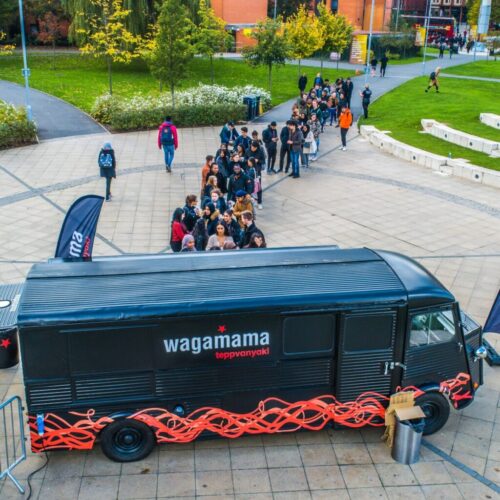 A long line of people wait outside a black food truck labeled wagamama teppanyaki parked on a paved area. Green grass and trees are visible in the background. Two flags with the wagamama logo stand near the truck. BAM Agency