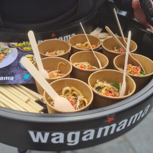 Small brown paper cups are filled with noodle stir-fry dishes, each with a wooden fork. They are arranged on a black tray with Wagamama printed on it. A Wagamama brochure is also visible on the tray. BAM Agency