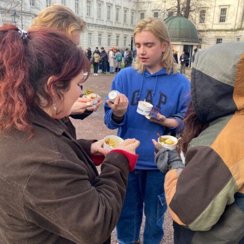 A group of four people is gathered outdoors, eating from small bowls. One person in a blue hoodie holds a container, possibly adding condiments. The background features a large building and other people milling about. It appears to be a chilly day. BAM Agency