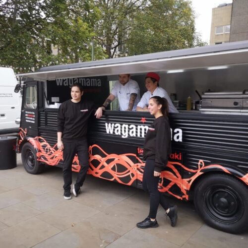 Food truck labeled wagamama with four people in black and white uniforms standing around it. The truck is parked on a paved area with trees and buildings in the background. BAM Agency