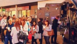 BAM Agency A diverse group of people stands in line before the wagamama noodle tour food truck, part of an engaging youth experiential campaign. The truck is parked on a lively street, with everyone eagerly holding cups and bags as they await their turn for some delicious delights. BAM Agency