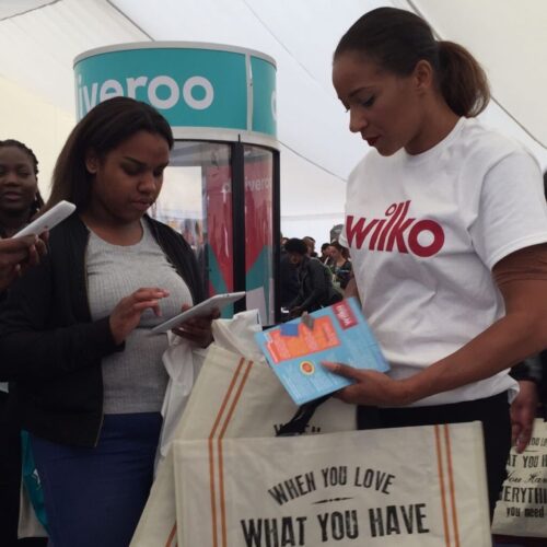 A woman wearing a wilko shirt hands out flyers and samples to a person looking at a smartphone. They are in a crowded tent with a Deliveroo booth in the background. BAM Agency