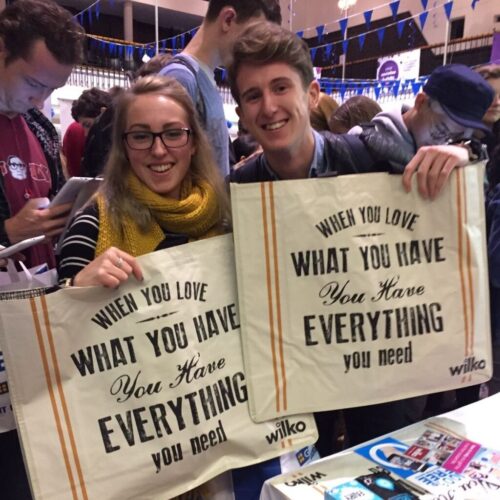 Two people smile while holding large tote bags with the text When you love what you have, you have everything you need. They appear to be at an indoor event with other attendees and blue and white bunting in the background. BAM Agency