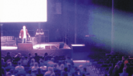BAM Agency A person stands on a stage at YMS 2016, addressing an audience in a dimly lit auditorium. The crowd, eager to learn about Youth Marketing Strategy, faces forward as bright lights create a spotlight effect over the speaker. BAM Agency