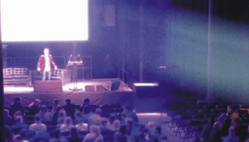 BAM Agency A person stands on a stage at YMS 2016, addressing an audience in a dimly lit auditorium. The crowd, eager to learn about Youth Marketing Strategy, faces forward as bright lights create a spotlight effect over the speaker. BAM Agency