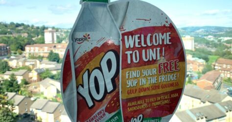 A window sticker with a Yop drink bottle illustration promotes a Welcome to Uni offer. It mentions finding a free Yop in the fridge and lists stores like Tesco and Sainsburys. The background shows a cityscape with trees and buildings under a blue sky. BAM Agency