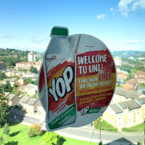 A window sticker with a Yop drink bottle illustration promotes a Welcome to Uni offer. It mentions finding a free Yop in the fridge and lists stores like Tesco and Sainsburys. The background shows a cityscape with trees and buildings under a blue sky. BAM Agency A window sticker with a Yop drink bottle illustration promotes a Welcome to Uni offer. It mentions finding a free Yop in the fridge and lists stores like Tesco and Sainsburys. The background shows a cityscape with trees and buildings under a blue sky. BAM Agency