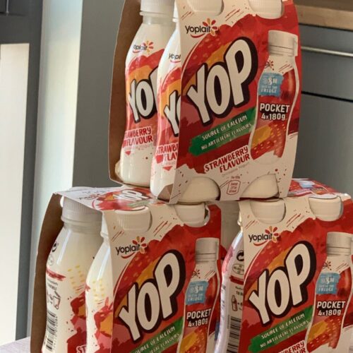 A stack of Yop yogurt drink bottles in strawberry flavor is arranged on a table. The packaging is predominantly white with red accents, and the Yop logo is prominently displayed. BAM Agency