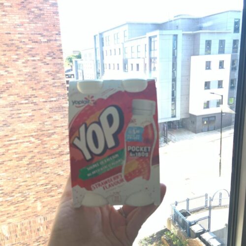 A hand holds two small bottles of Yop strawberry-flavored drink against the backdrop of a cityscape with modern buildings and a partly cloudy sky. BAM Agency