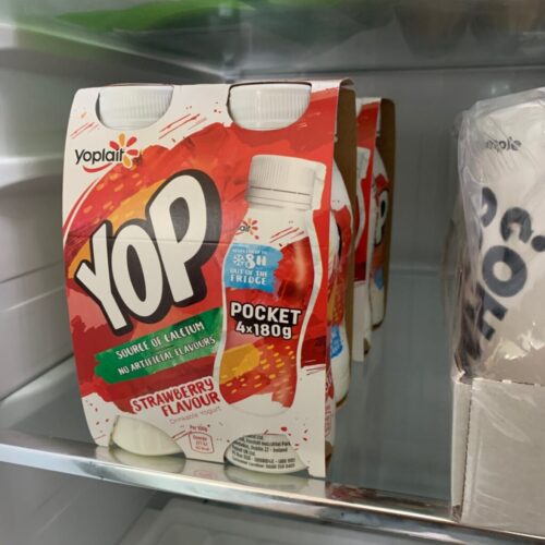 Yogurt drink bottles labeled YOP in strawberry flavor are displayed in a refrigerator. The packaging highlights source of calcium and no artificial flavors with 4x180g packs. Another branded box is partially visible to the right. BAM Agency